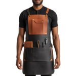 Bicolore Stratega — Men's Jet Black Waxed Canvas Body Tan Cognac Genuine Full-Grain Cowhide Leather Two-Tone Contrast Heavy-Duty Adjustable Black Cross-Back Strap Antique Brass Buckle Large Tan Leather Chest Flap Pocket Multiple Side Tool Loop Knife Holder Wide Lower Canvas Panel Gathered Waistband Chef Barber Blacksmith Carpenter Craftsman Bold Two-Tone Professional Workshop Utility Apron